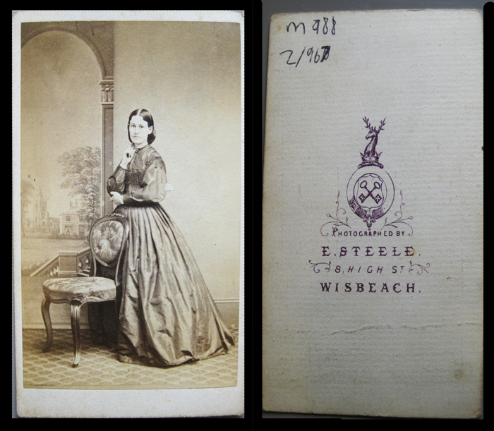 Carte de visite, 1860s, unknown lady in criniline, painted backcloth with archway and country house and diamond pattern carpet. Reverse has heraldic design with garter cross keys, stag's head and crown. Town name spelled "Wisbeach".