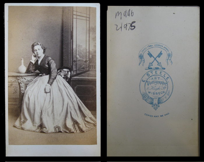 Carte de visite, 1860s, unknown lady in criniline, painted backcloth with open window and garden, with ornate table and bottle. Reverse has heraldic design with garter cross keys and Latin motto. Town name spelled "Wisbech".